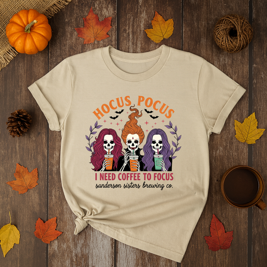 Halloween t-shirt with Sanderson Sisters skeletons holding coffee cups and text “Hocus Pocus I Need Coffee to Focus” – spooky coffee lover tee.
