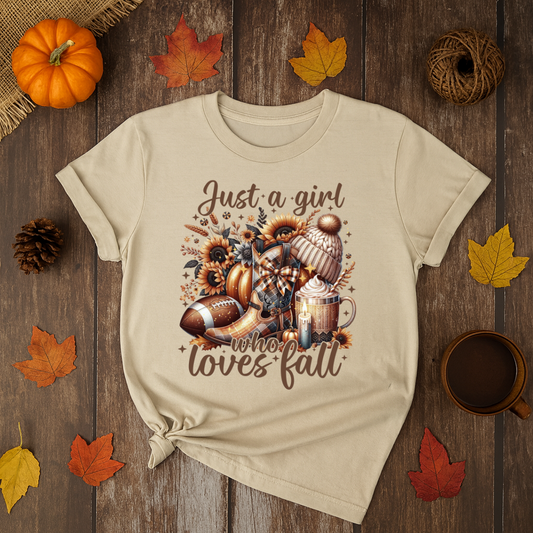 Beige t-shirt with 'Just a girl who loves fall' design on a wooden surface with autumn decorations.